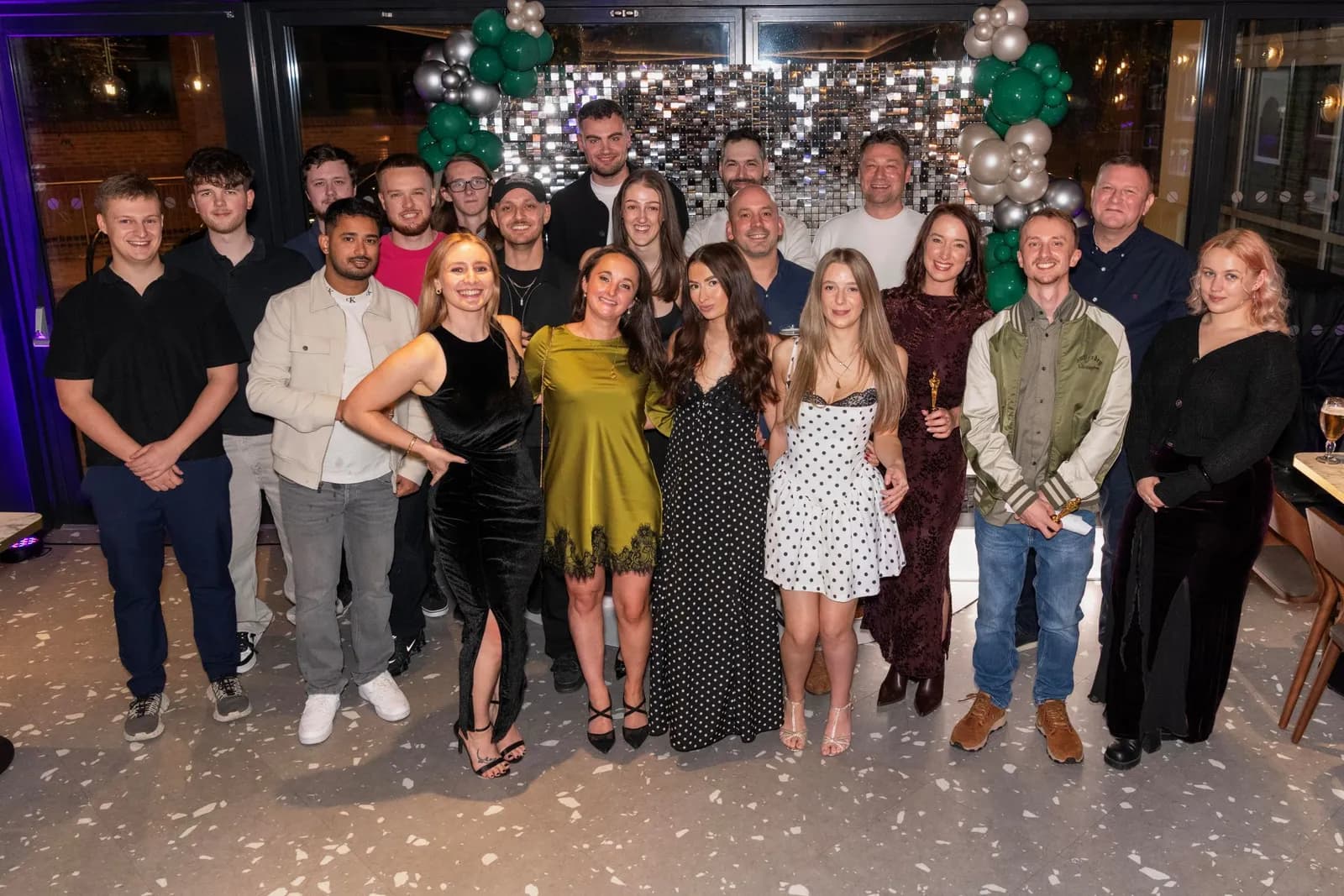A large group of people are gathered for a photo, with men mostly in casual button-down shirts and women in dresses, standing in front of a shiny wall decorated with green and silver balloons.