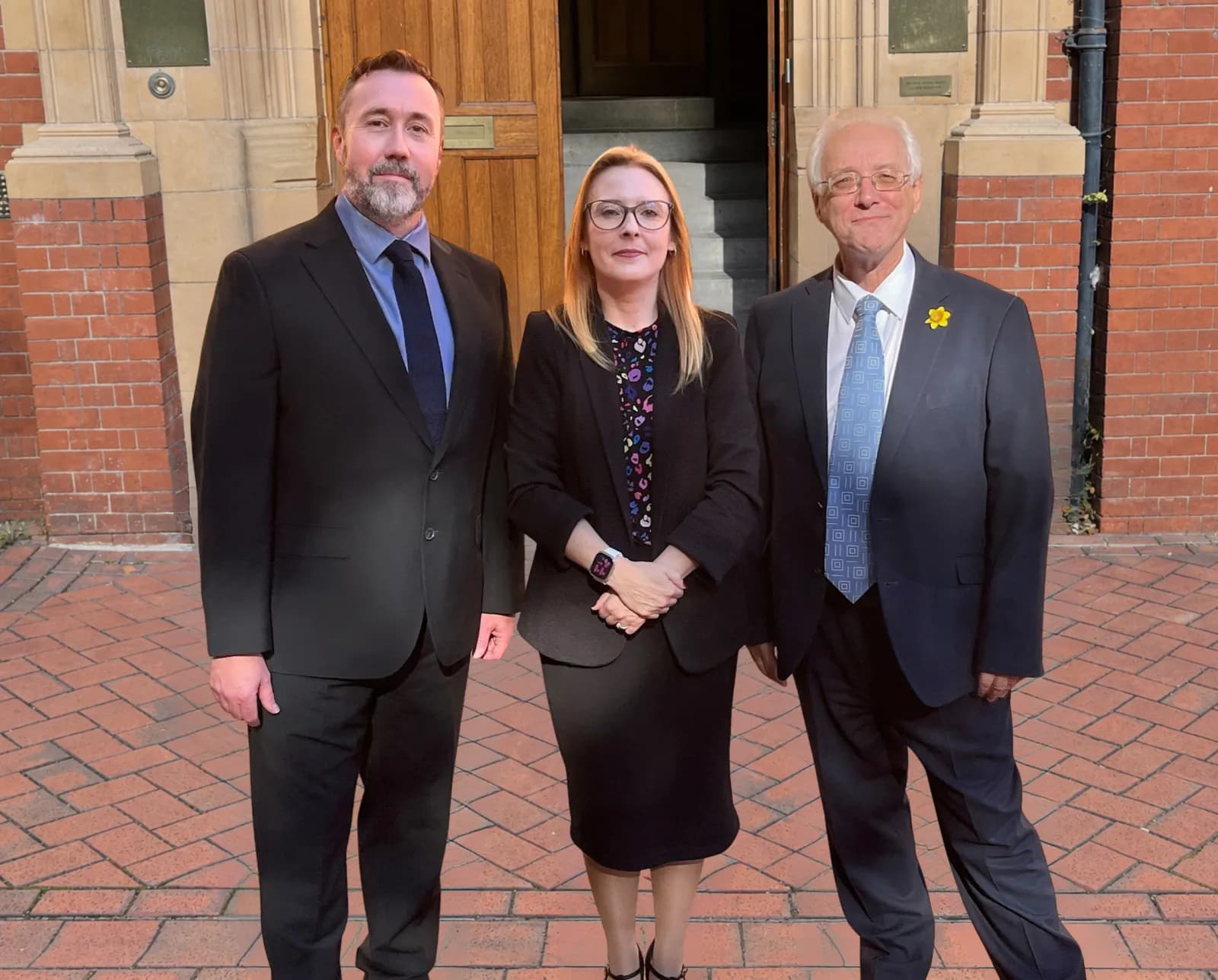 Three people, two men and one woman, in business attire stand outdoors in front of a brick building with a wooden door.