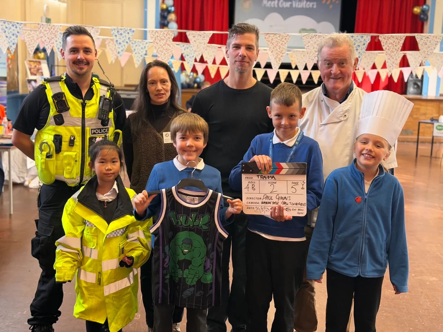 A group of adults and children pose for a photo in a decorated hall, with a police officer, a chef, and children dressed in different professional outfits.