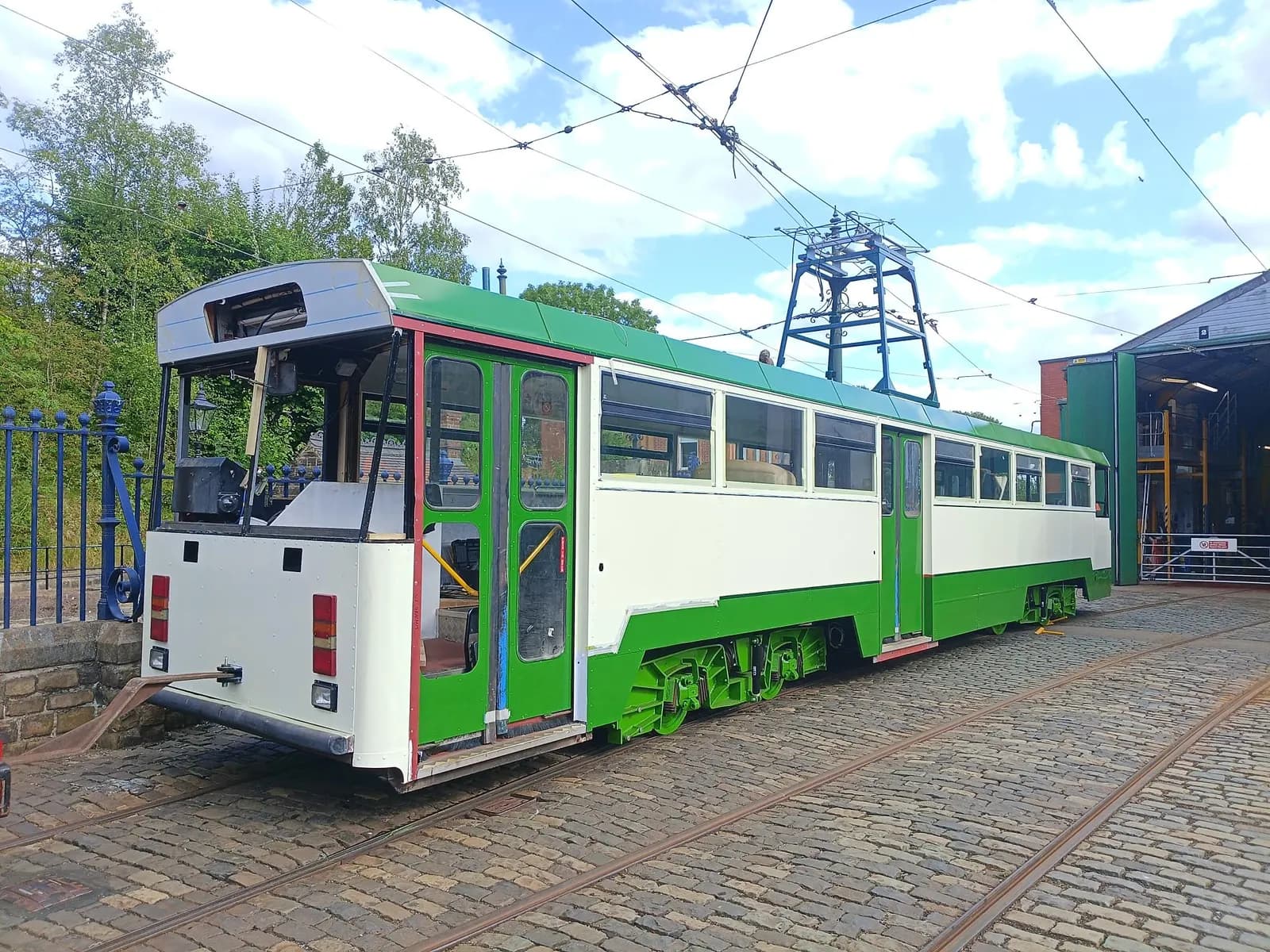 A long, green and white tram car sits on cobblestone tracks under a bright, cloudy sky, with a building and trees in the background.